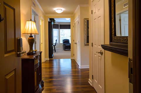 View down a well-lit hardwood-floored hallway with a lamp on a console table, doors and a mirror on the right, leading into a sunlit sitting area with a chair and window.