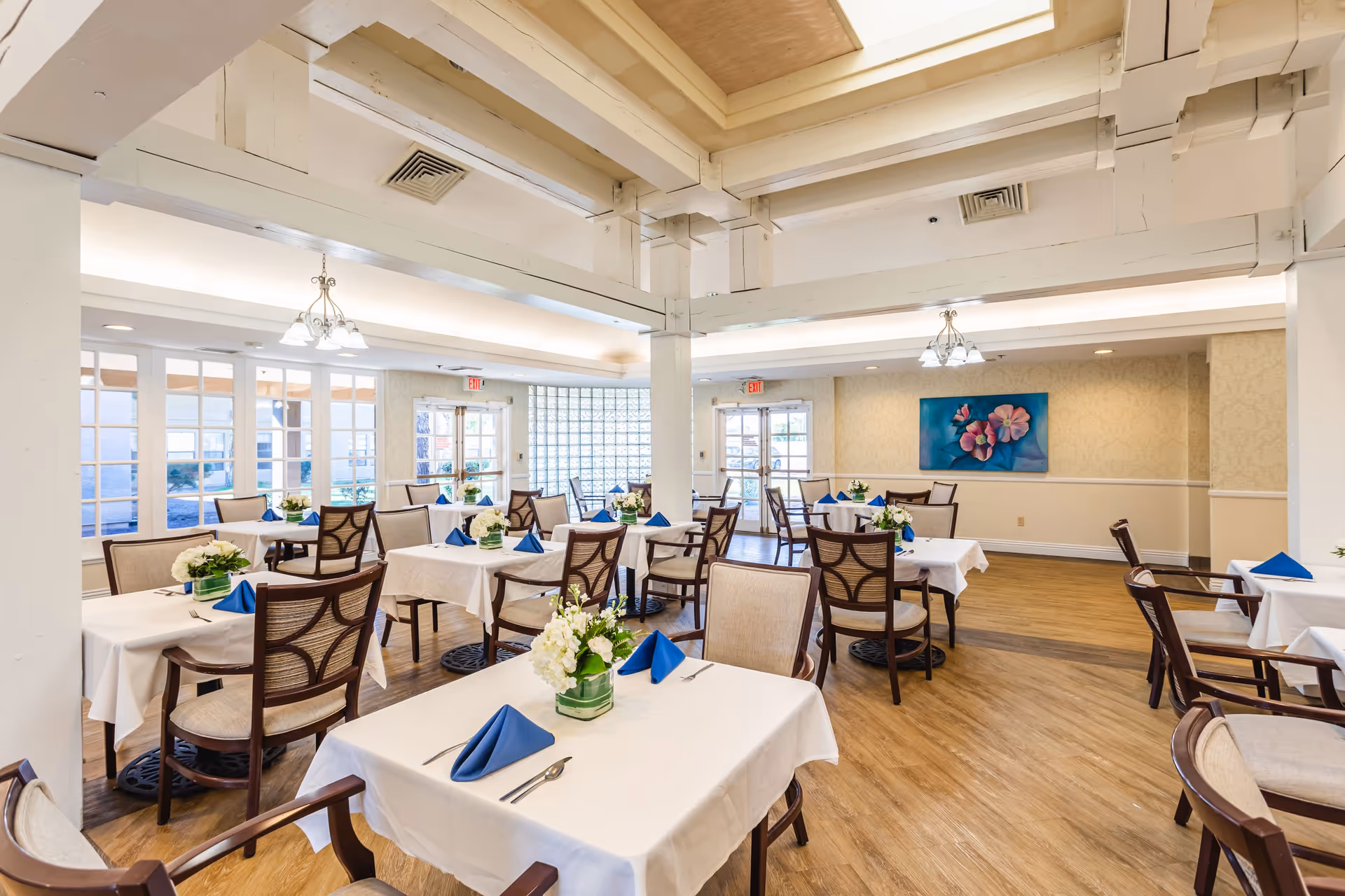 A bright and spacious dining room with multiple tables covered in white tablecloths, each set with blue folded napkins, silverware, and floral centerpieces. The room has wooden chairs with cushioned seats, large windows allowing natural light, and a painting of pink flowers on a blue background on the far wall. The ceiling features exposed beams and hanging light fixtures.