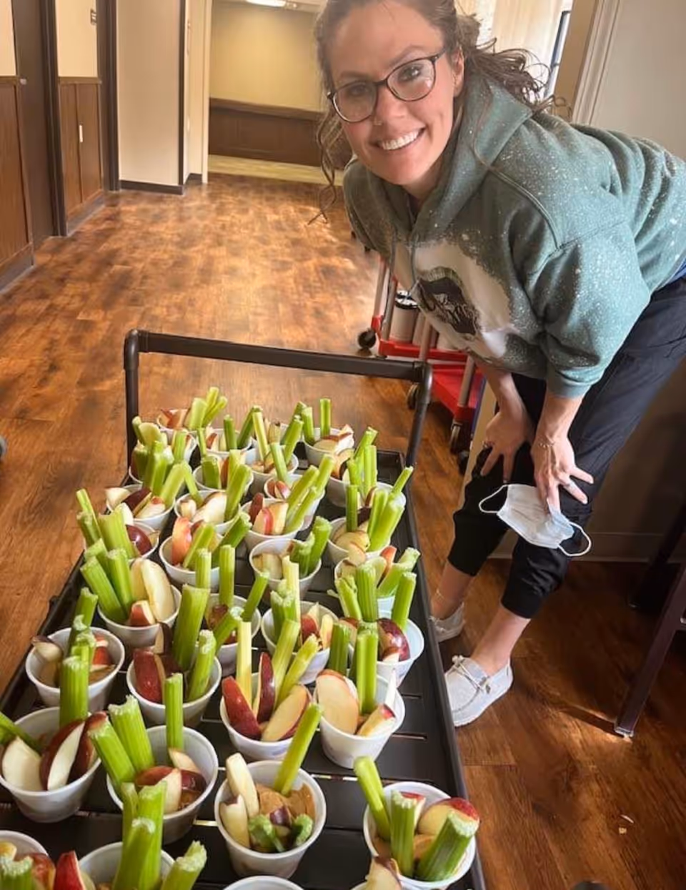 A woman wearing glasses and a green hoodie is smiling and leaning over a cart filled with small cups containing celery sticks and apple slices. She is holding a face mask in one hand and is in a room with wooden floors and wood-paneled walls.