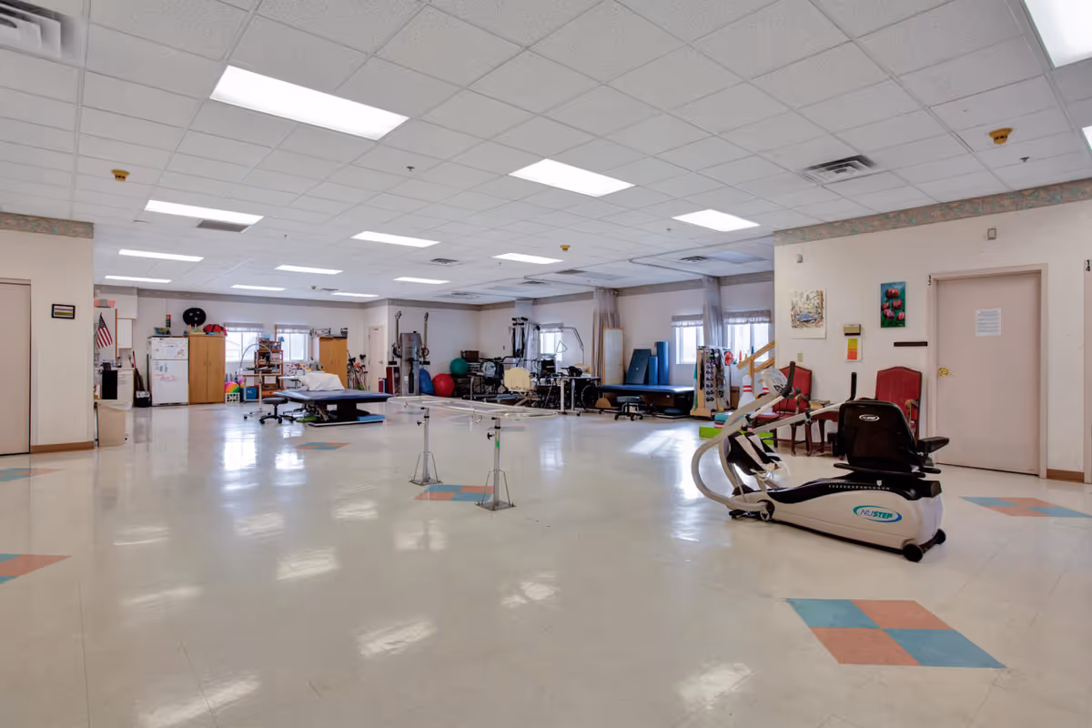 Large bright physical therapy/recreation room with exercise equipment, therapy tables, and chairs.