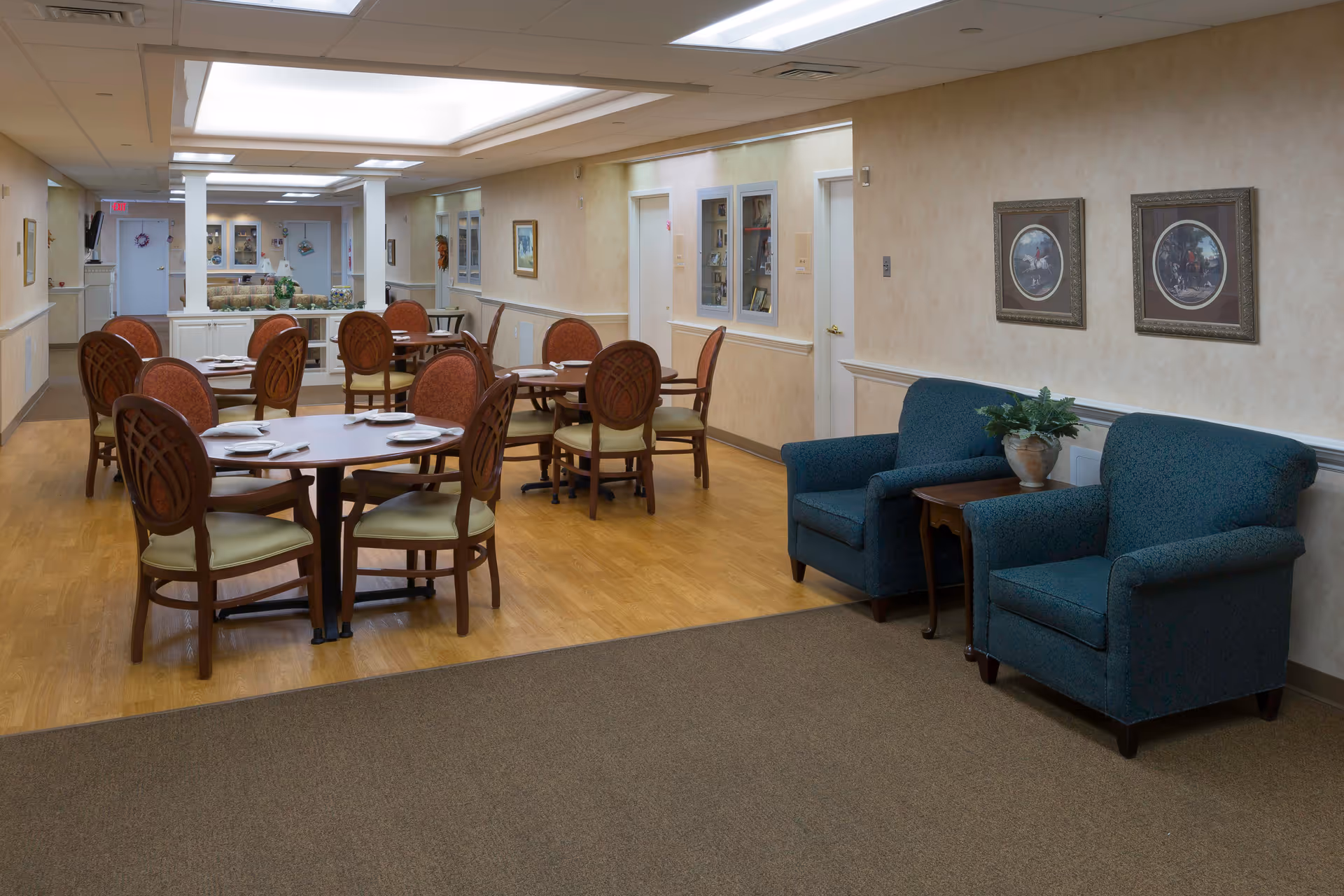 Interior view of a senior living facility dining area with several round wooden tables and chairs set with plates and napkins. To the right, there are two blue upholstered armchairs with a small wooden table and a potted plant between them. The walls are beige with framed artwork, and the floor is a combination of wood and carpet. The space is well-lit with ceiling lights and has a cozy, welcoming atmosphere.