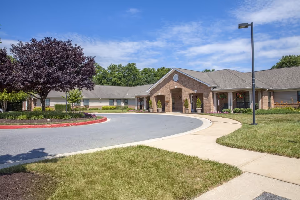 Front exterior view of a single-story senior living facility building with a circular driveway, landscaped greenery including a purple-leafed tree, and a clear blue sky with some clouds.