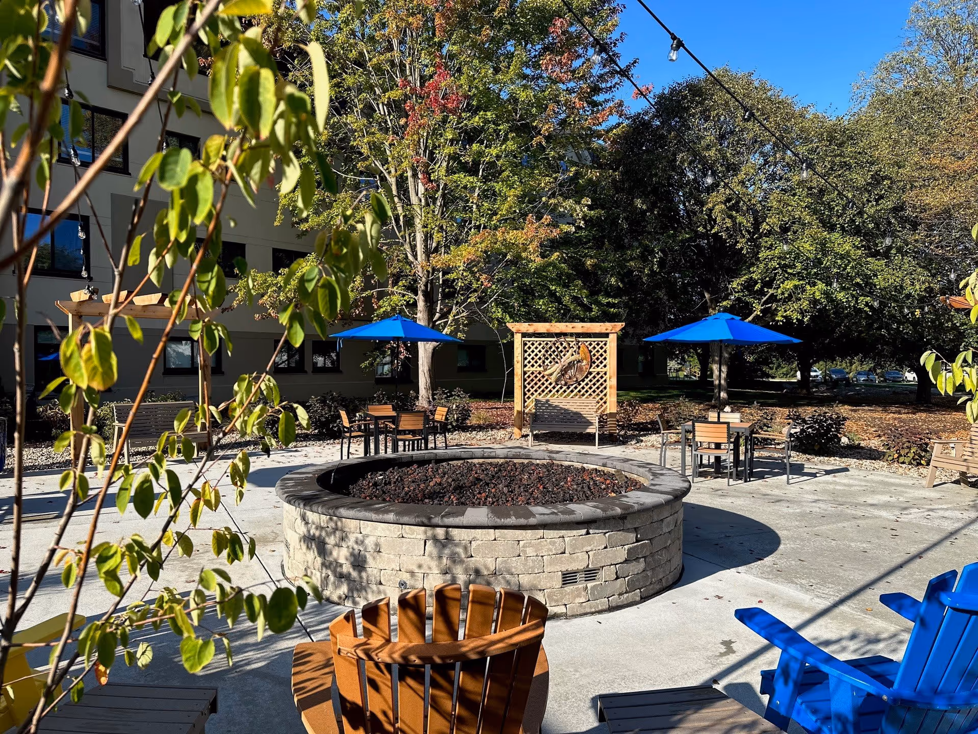 Outdoor patio area at Westminster Village featuring a circular stone fire pit surrounded by wooden and blue Adirondack chairs. There are tables with blue umbrellas and a wooden lattice structure with a decorative item hanging on it. Trees with green and some autumn-colored leaves surround the area under a clear blue sky.