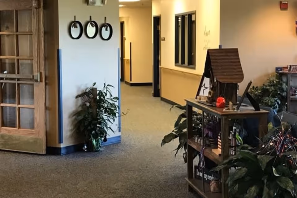 Interior hallway of a senior living facility with beige walls and carpeted floor. There is a wooden door with glass panels on the left, three round mirrors on the wall, and several green plants placed around. A wooden shelf with decorative items and a small house model is on the right side. The hallway extends into the background with doors and windows along the walls.