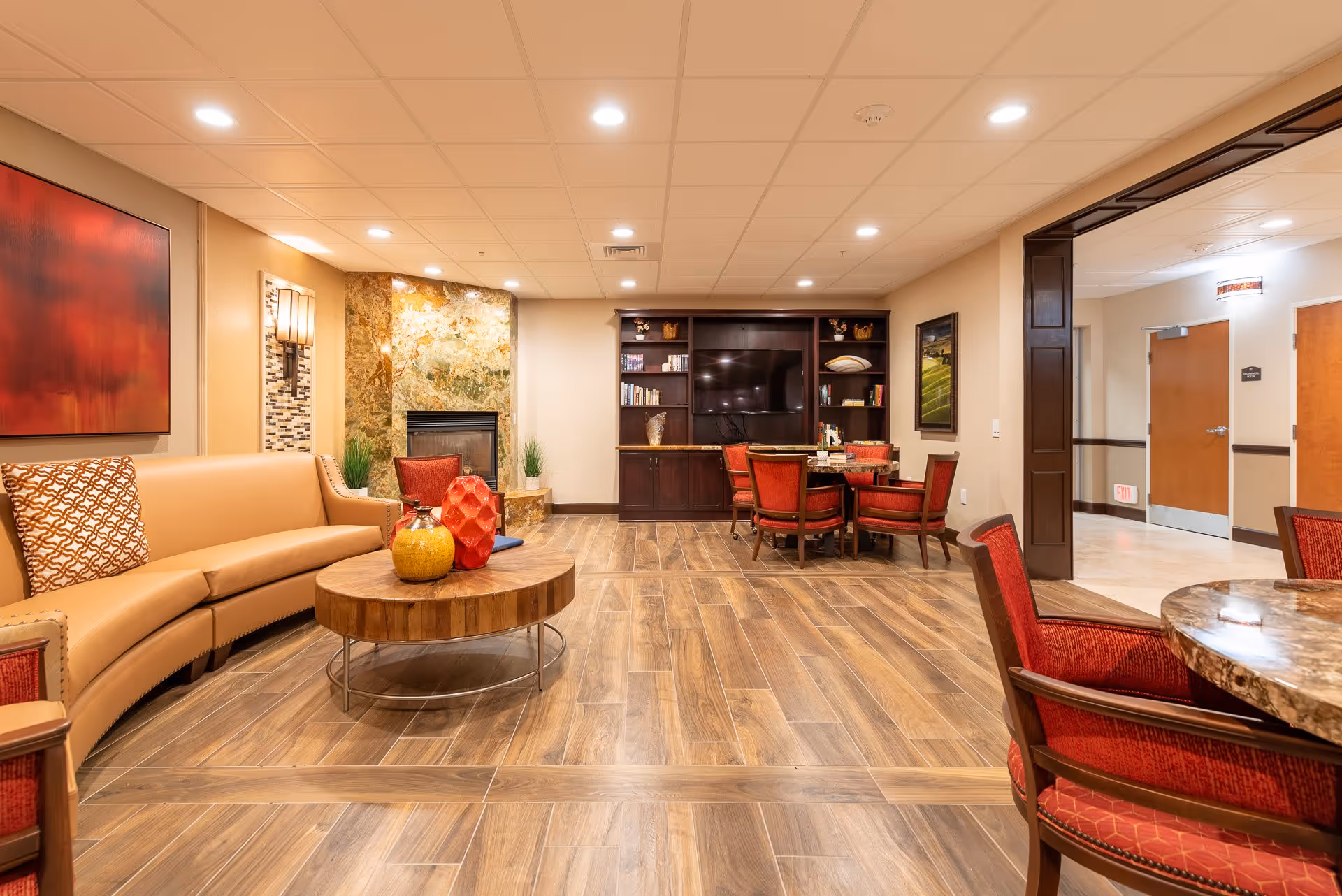 A cozy senior living common area with a tan curved sofa, a round wooden coffee table with decorative vases, red cushioned chairs around a round table, a fireplace with a stone surround, a large flat-screen TV mounted on a dark wooden entertainment center with shelves holding books and decor, warm lighting, and wood-look tile flooring.
