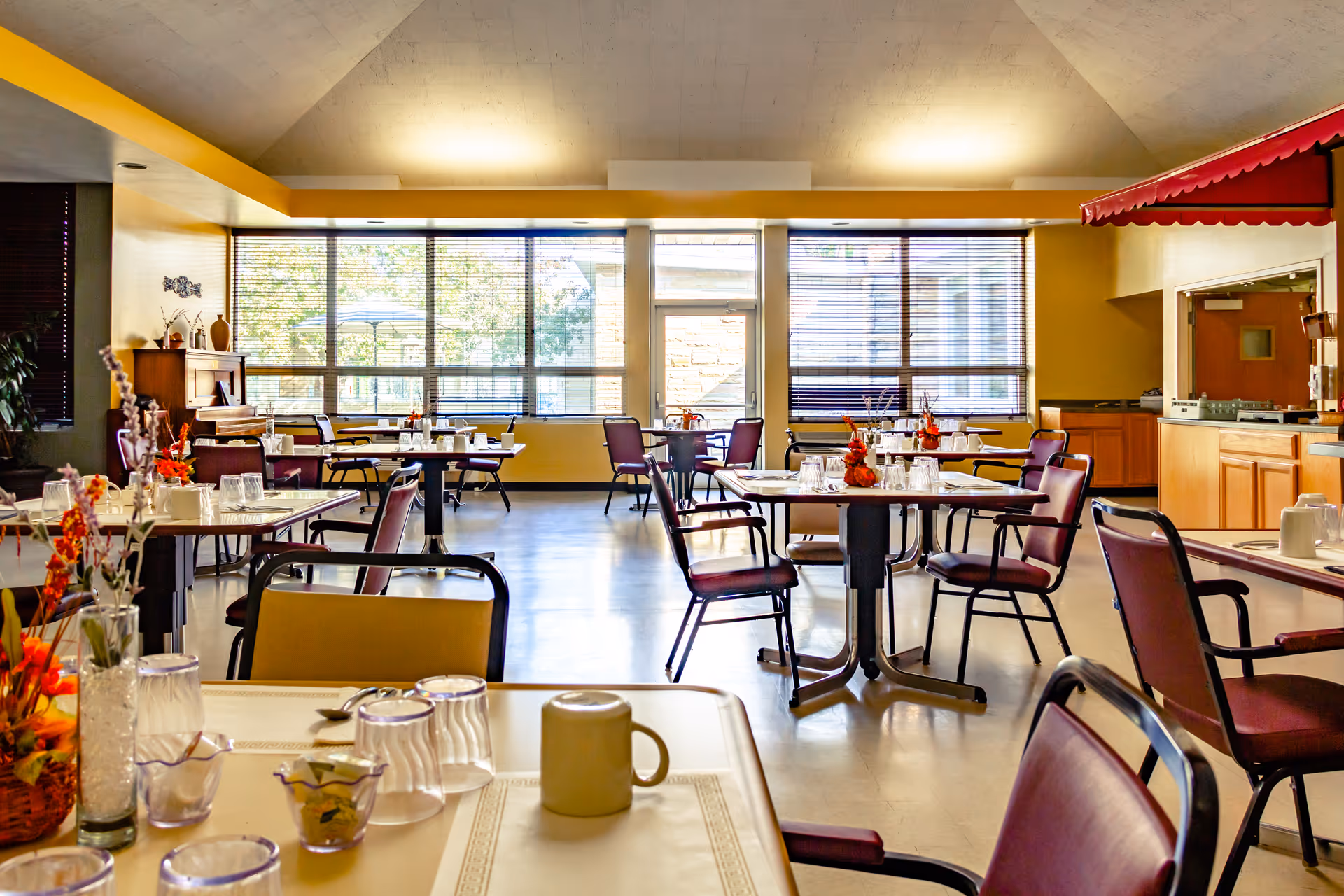 A bright dining room in a senior living facility with multiple tables and chairs arranged neatly. Each table is set with glasses, mugs, and small floral centerpieces. Large windows with blinds allow natural light to fill the room. There is a counter area on the right side with a red awning above it.