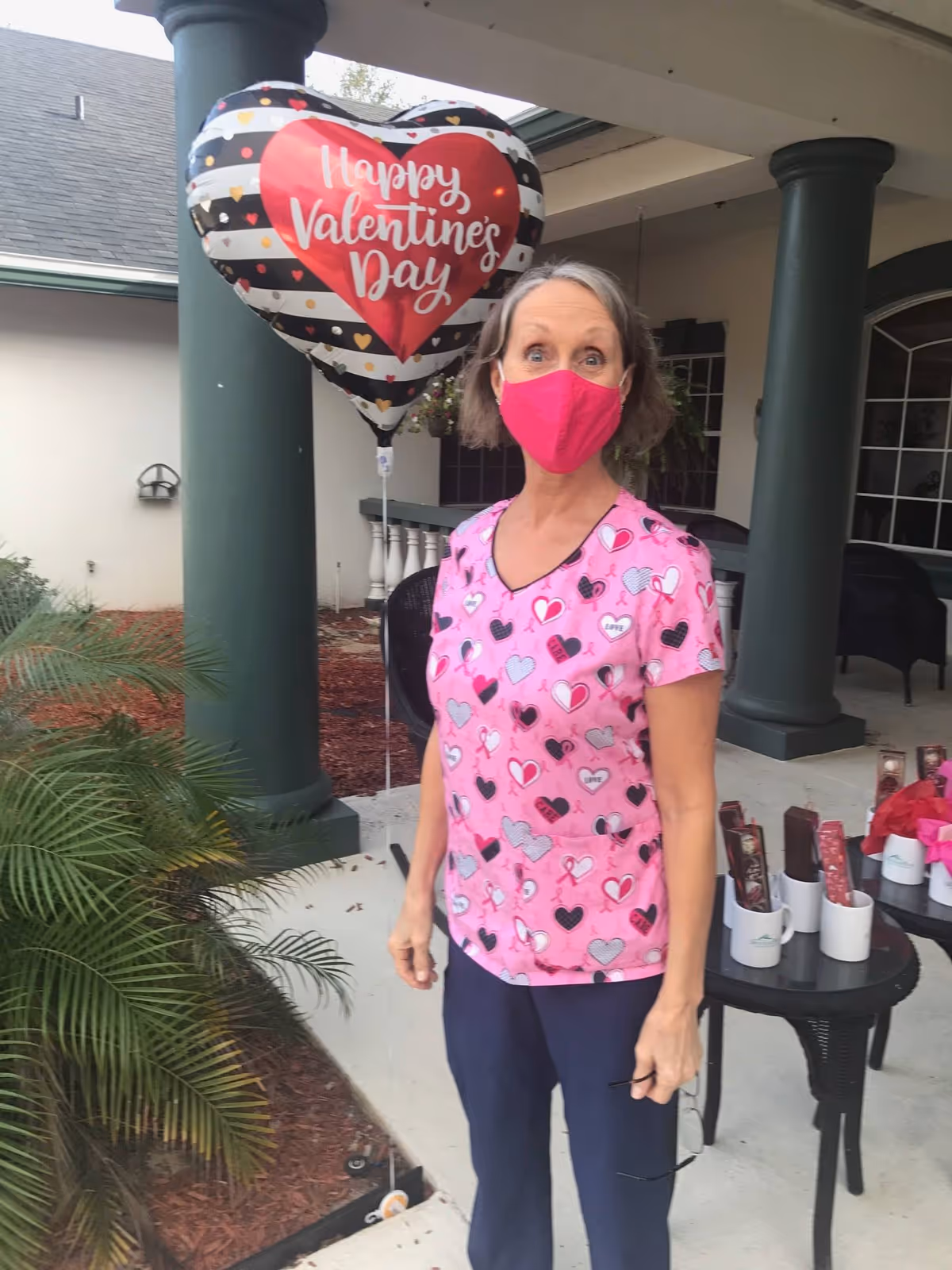 A woman wearing a pink heart-patterned scrub top and a bright pink face mask stands outdoors near a green pillar. Behind her, there is a heart-shaped balloon with the text 'Happy Valentine's Day'. There are plants and a table with various items on it in the background.