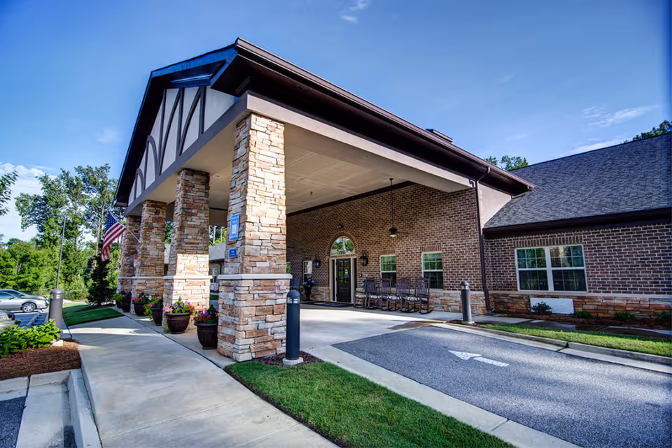 Entrance of a senior living facility with a covered drop-off area supported by stone pillars, an American flag, several rocking chairs near the door, and a well-maintained sidewalk and landscaping under a clear blue sky.