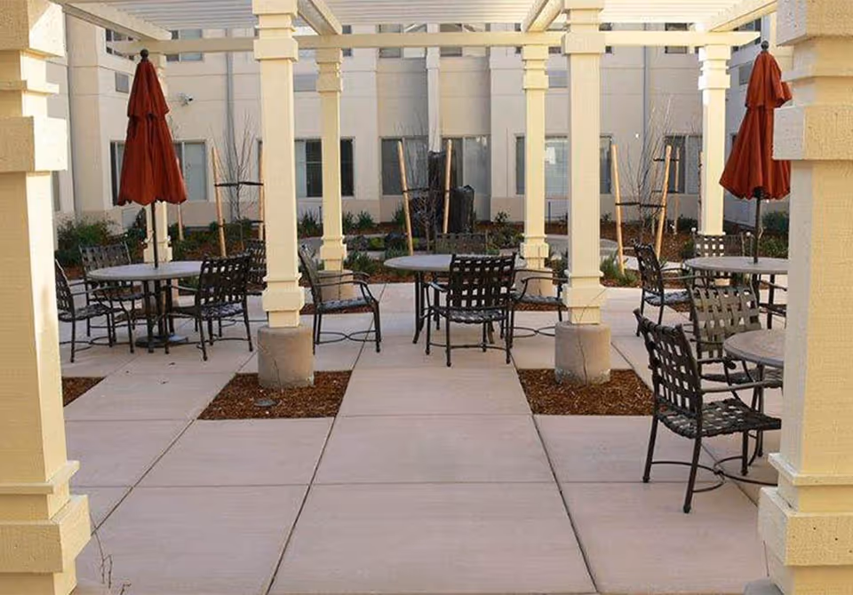 Outdoor patio area with multiple round tables and metal chairs under a pergola structure. Each table has a closed red umbrella. The area is paved with concrete and surrounded by garden beds with small plants and trees. The background shows the exterior walls and windows of a building.