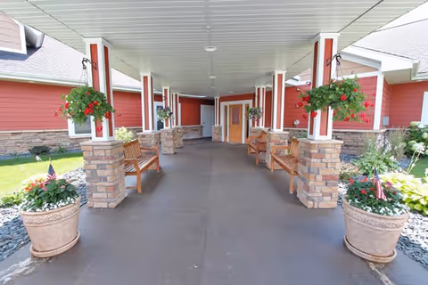 Covered outdoor walkway with stone pillars and wooden benches on both sides, hanging flower baskets attached to the pillars, and large potted plants with flowers and small American flags at the entrance. The building has red siding and a light-colored roof.