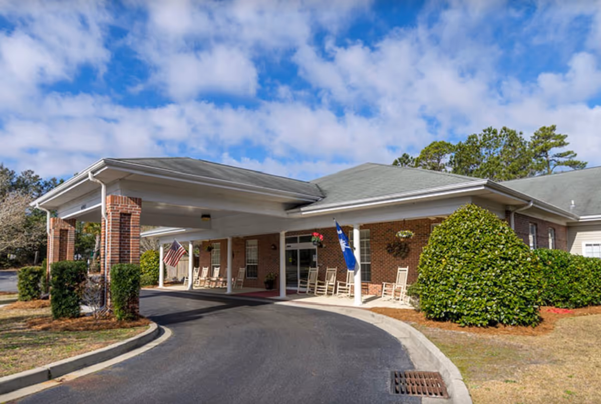 Exterior view of The Legacy of North Myrtle Beach facility showing a covered entrance with brick pillars, several rocking chairs on the porch, American and South Carolina state flags, surrounded by greenery and trees under a partly cloudy sky.