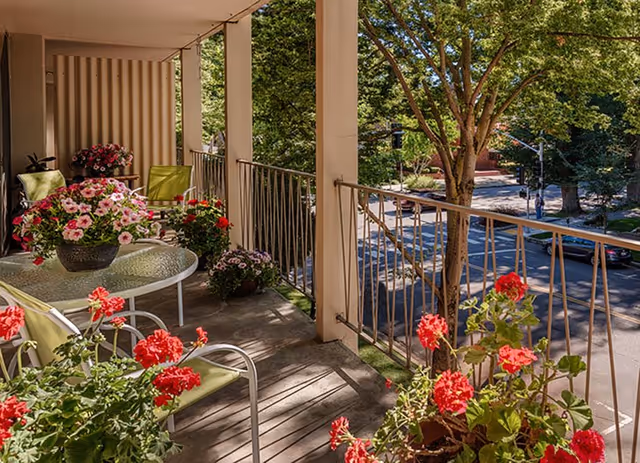 A balcony with green cushioned chairs and a glass-top table surrounded by vibrant potted flowers, overlooking a tree-lined street with cars and pedestrians.
