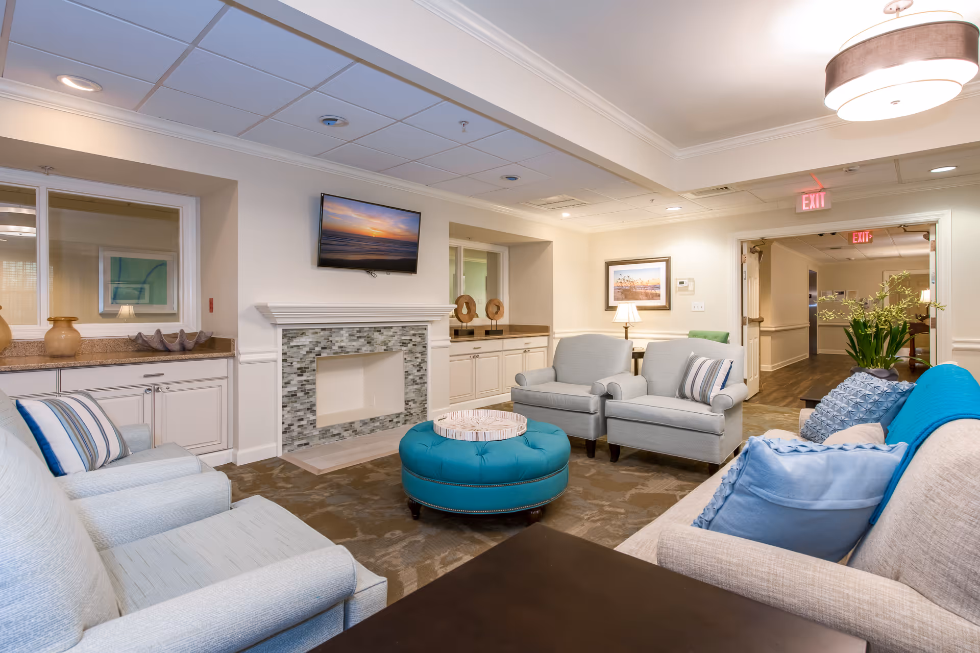 Bright communal lounge with sofas, armchairs, a teal round ottoman, fireplace, and a wall-mounted TV.