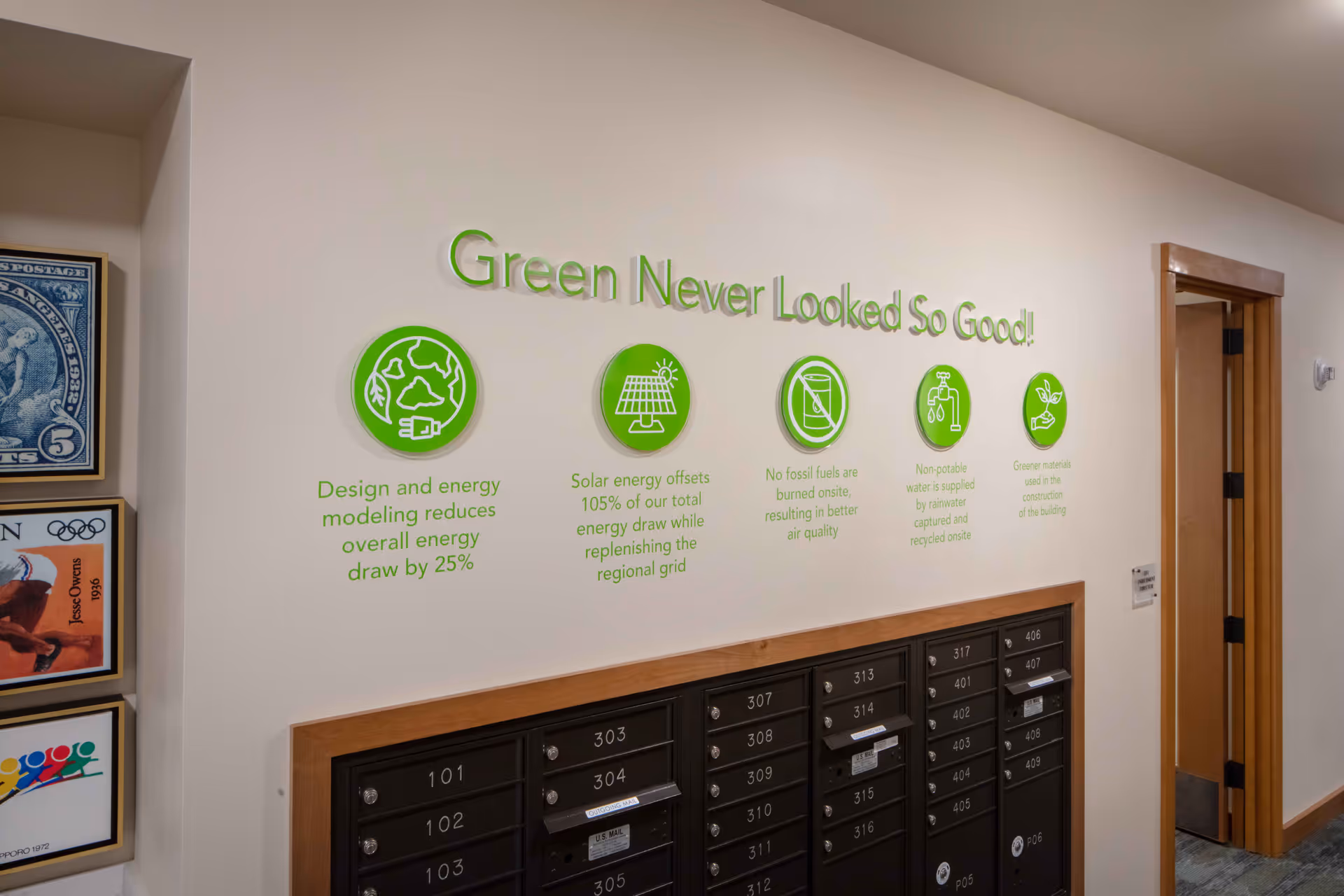 Interior hallway wall with a green-themed environmental message titled 'Green Never Looked So Good!' above a set of black mailboxes. The message highlights energy and sustainability features such as energy modeling reducing energy draw by 25%, solar energy offsetting 105% of total energy draw, no fossil fuels burned onsite, use of rainwater for non-potable water, and greener materials used in construction. To the left, framed pictures are partially visible, and to the right, a wooden door is open.