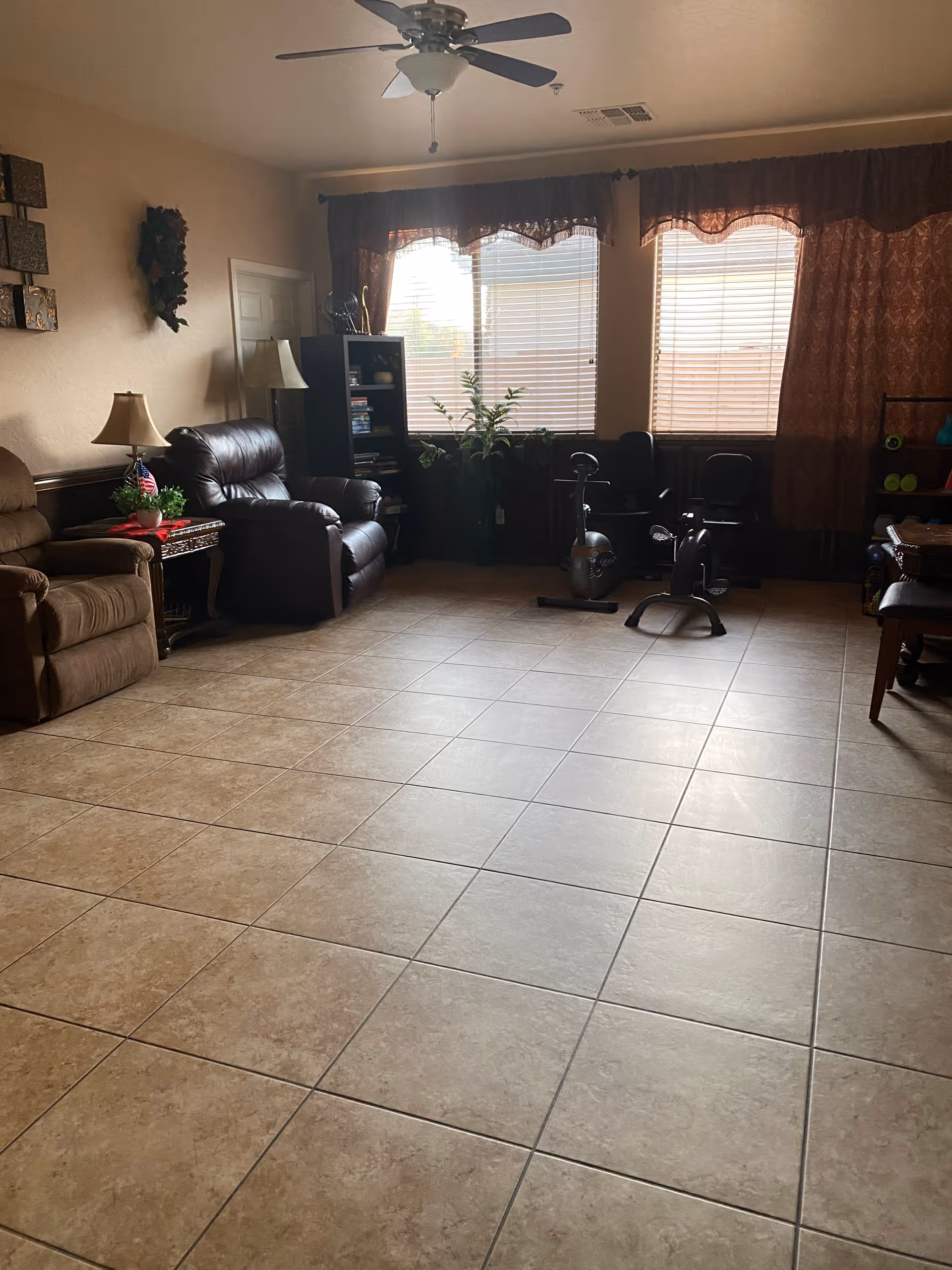 A living room with tiled floor, two recliner chairs, a side table with a lamp and a small plant, a bookshelf with books and decorative items, two exercise bikes near windows with blinds and brown curtains, a ceiling fan, and some wall decorations.