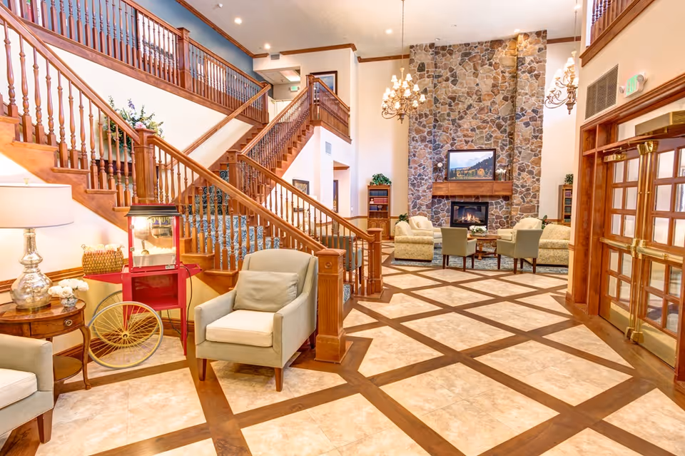 Bright, elegant lobby with a large stone fireplace, multiple seating areas, a wooden staircase, and patterned tile flooring.