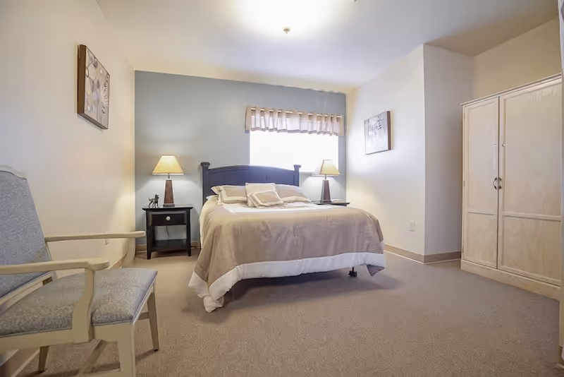 Well-lit bedroom with a centrally placed bed, two bedside tables with lamps, a wardrobe, and a chair.