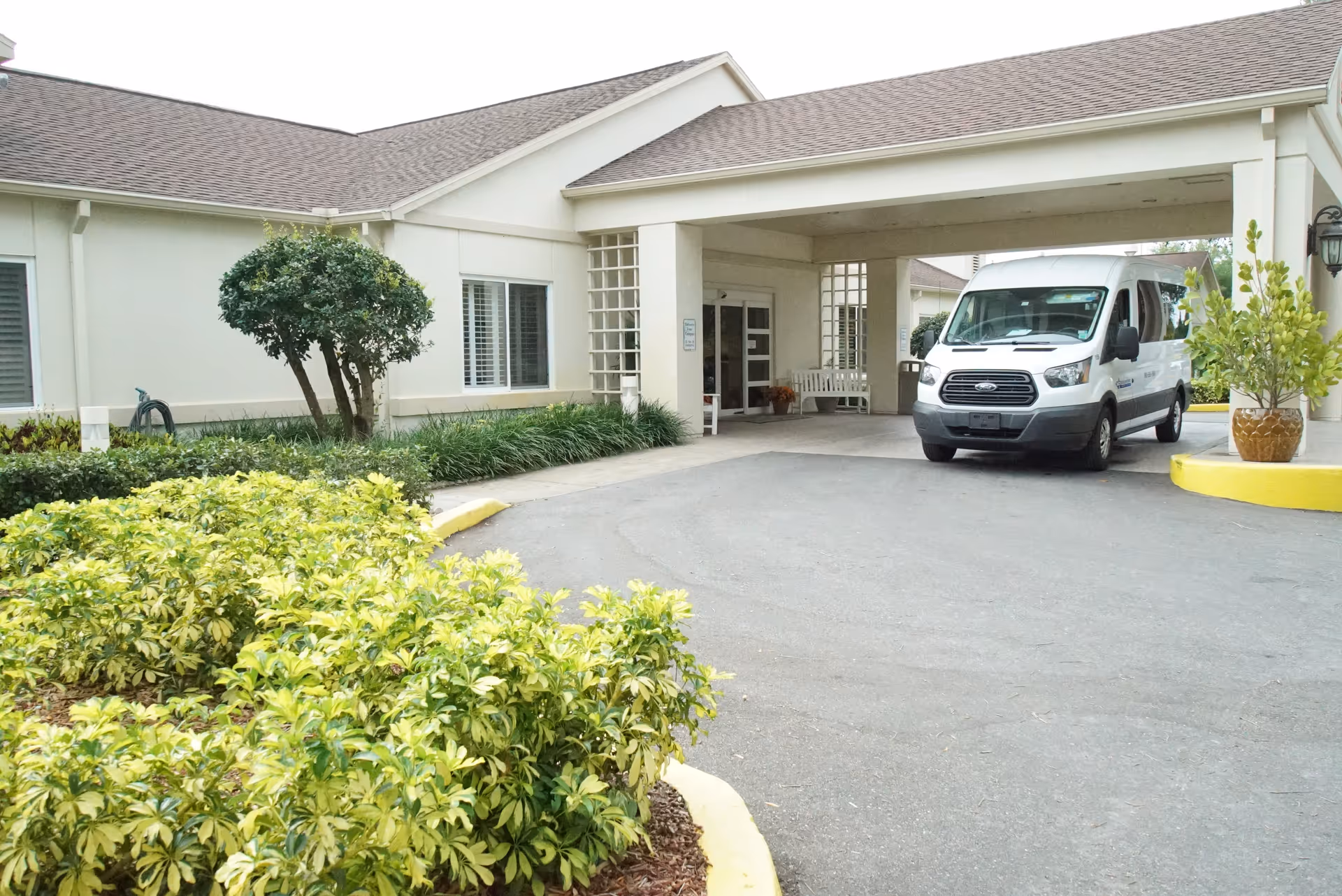 Front entrance of a senior living facility with a covered drop-off, a white shuttle van parked under the porte-cochere, and landscaped shrubs in front.