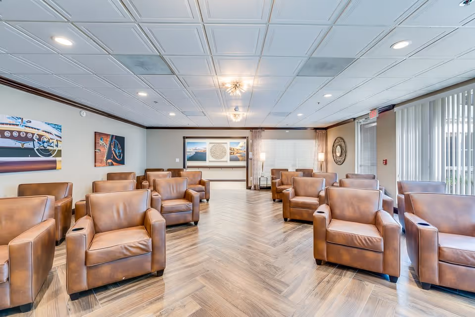 A spacious senior living facility common area with multiple brown leather armchairs arranged in rows on a wooden herringbone floor. The room has a white paneled ceiling with recessed lighting and modern light fixtures. Large windows with vertical blinds allow natural light to enter. The walls are decorated with framed artwork and a round decorative mirror.