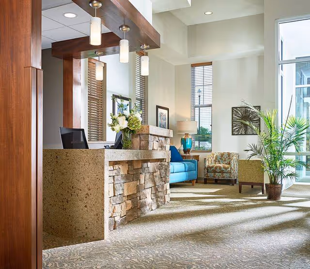 A bright and welcoming senior living facility lobby with a stone and concrete reception desk, pendant lights, a blue sofa, patterned armchairs, a table lamp, framed artwork, large windows with blinds, and a potted plant.