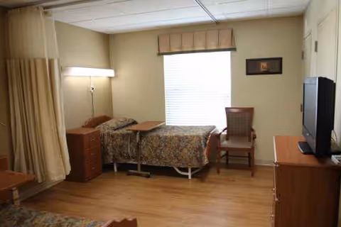 A shared senior living bedroom with two beds, a nightstand, chair, dresser, and TV.