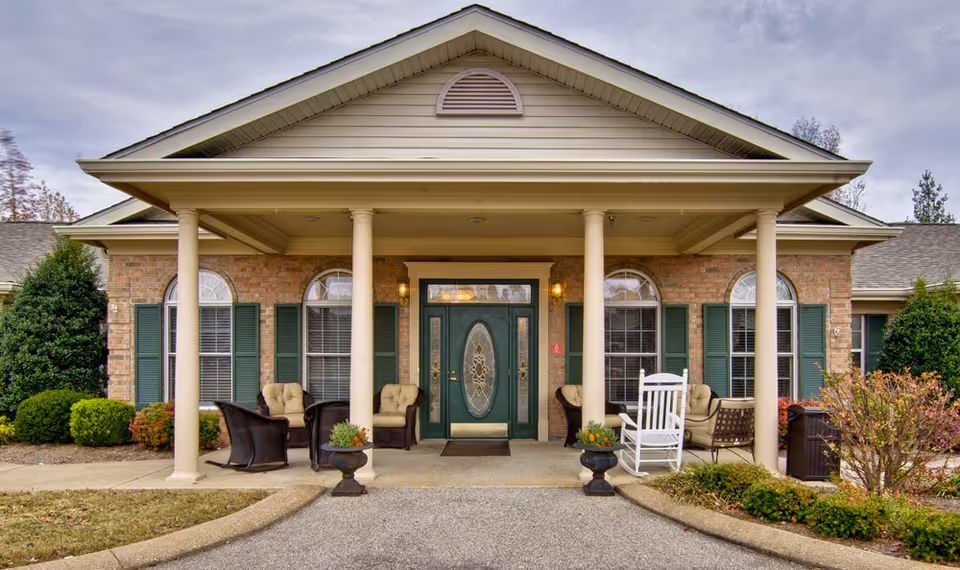 Front entrance of a senior living facility with a covered porch supported by four columns. The building has a brick facade with green shutters on the windows. There are cushioned chairs and a white rocking chair arranged on the porch, along with two planters with flowers near the entrance. The sky is overcast.
