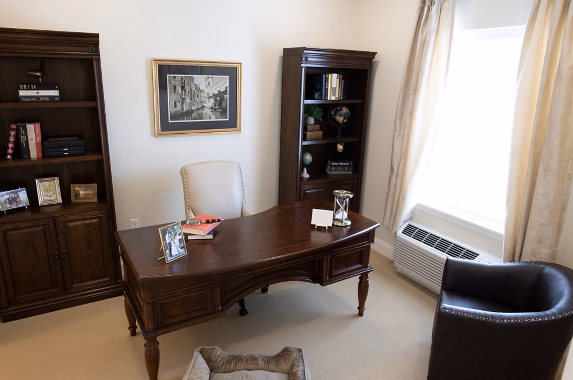 A furnished interior office with a wooden desk, upholstered chair, bookshelves, framed art, and a curtained window.