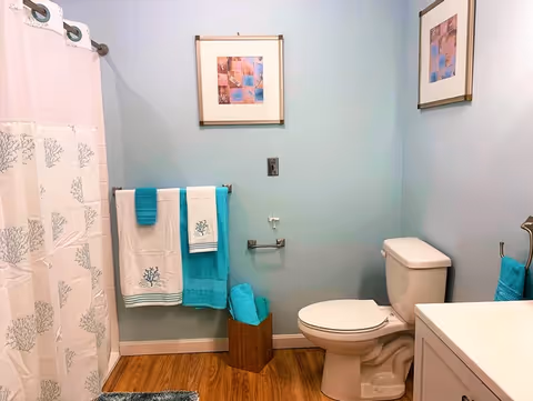 A light-blue walled bathroom with a toilet, vanity, shower curtain, and turquoise-accented towels and decor.
