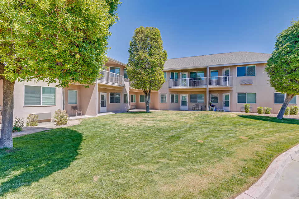 Outdoor view of a senior living facility courtyard with a well-maintained lawn, several trees, and a two-story building with balconies and windows under a clear blue sky.