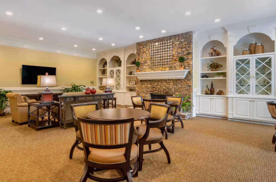 Community lounge with round tables and chairs, built-in shelving, a stone fireplace and a wall-mounted TV.