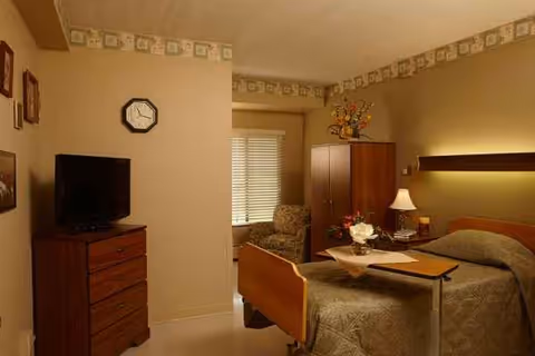 Cozy senior living bedroom with bed, dresser, TV, table, lamp, and floral decor.