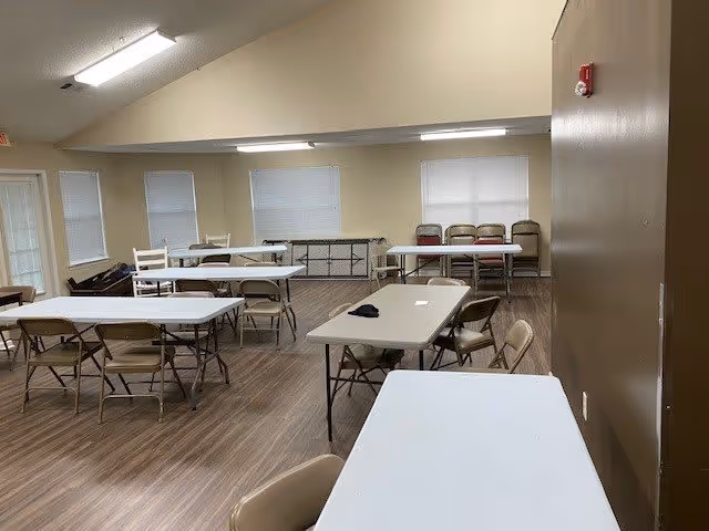 A spacious room with several rectangular folding tables and folding chairs arranged around them. The room has beige walls, multiple windows with closed blinds, and a wood-patterned floor. Fluorescent lights are mounted on the ceiling, and there is a fire alarm on one wall.