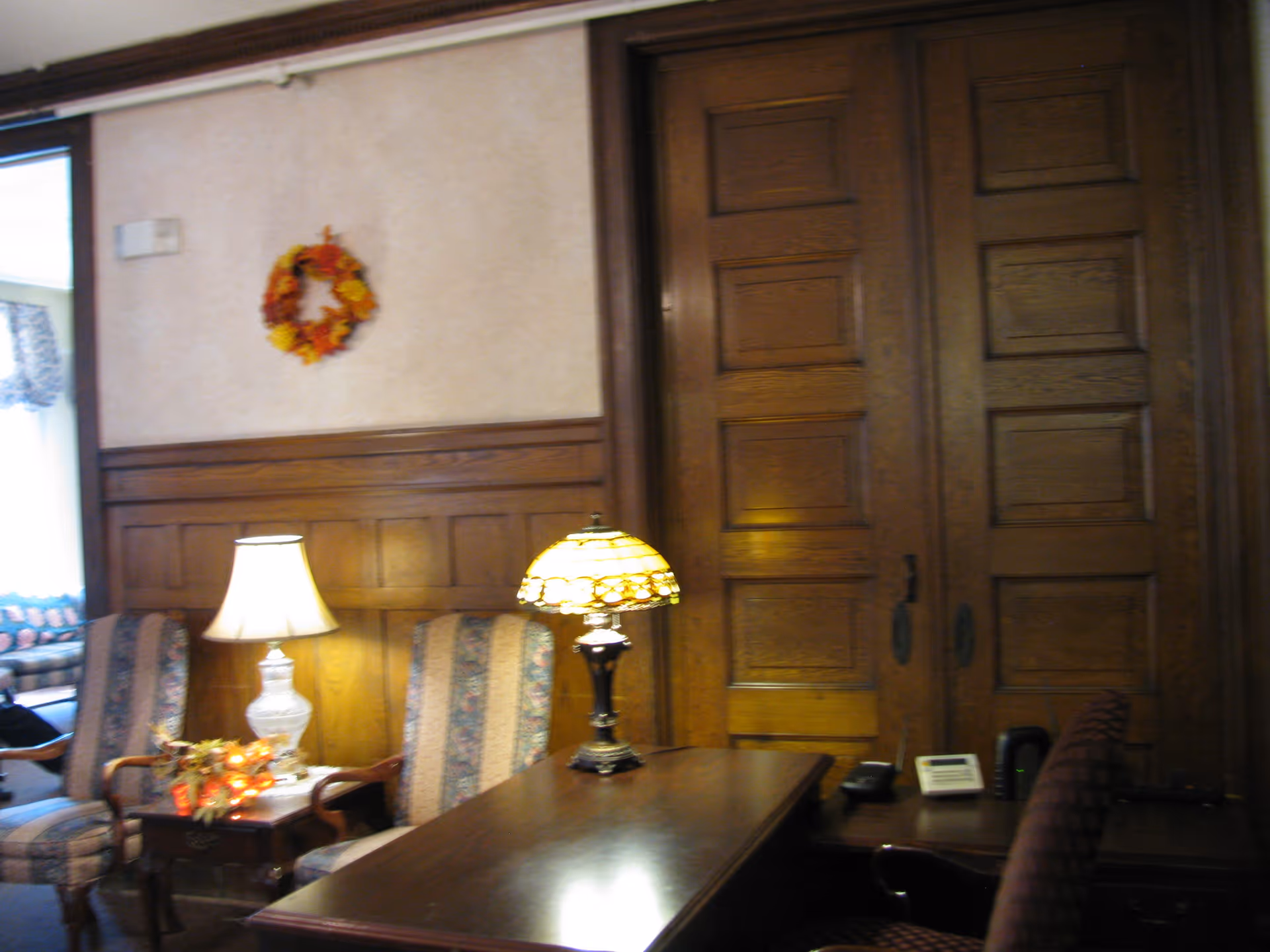 A cozy interior room with wooden paneling on the walls and large wooden double doors. There is a wooden desk with a Tiffany-style lamp on it, a telephone, and a chair with a patterned fabric. Two upholstered armchairs with floral and striped patterns sit next to a small side table with a white lamp and a decorative autumn wreath hanging on the wall above.