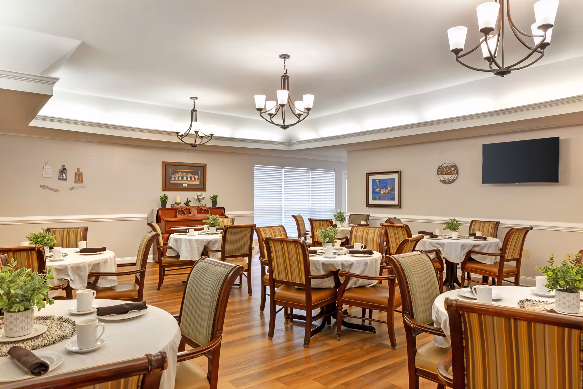 Bright dining room with round tables set for meals, wooden chairs, chandeliers, and a wall-mounted TV.