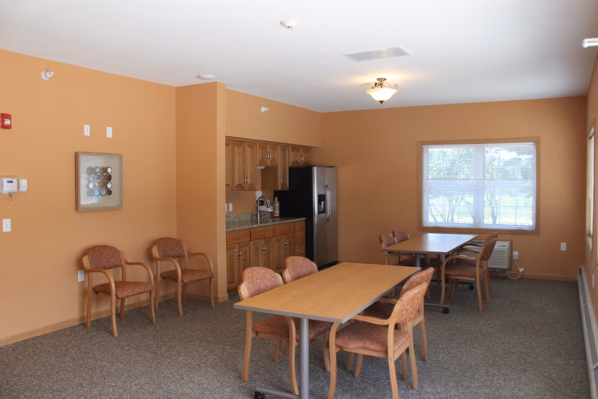 Communal dining area with tables, chairs, and a kitchenette against warm-colored walls.