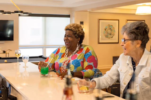 Two elderly women sitting at a bar counter in a well-lit room, enjoying colorful drinks and engaging in conversation. One woman is wearing a colorful patterned top and white necklace, while the other is wearing glasses and a white jacket. The background includes a window with blinds, a wall-mounted TV, and framed artwork.