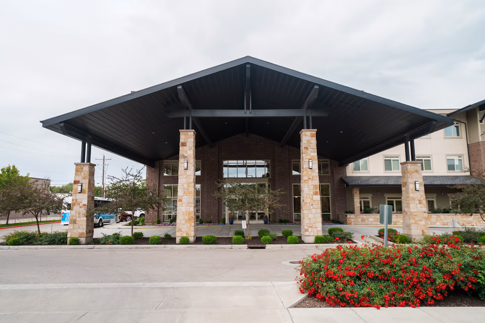 Front exterior view of Creekside Assisted & Senior Living facility featuring a large covered entrance supported by stone pillars, landscaped bushes, and red flowers in the foreground under a cloudy sky.