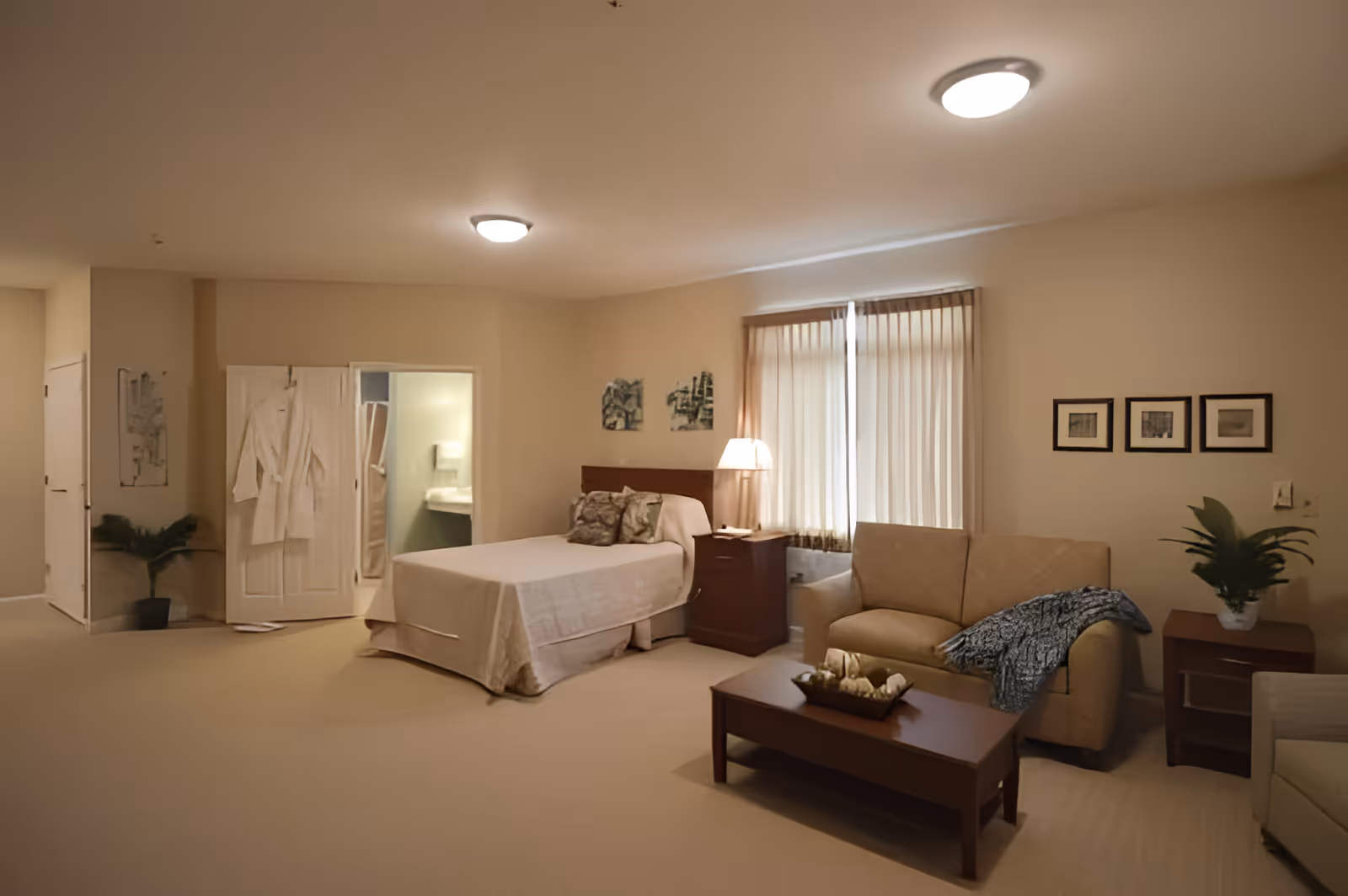 A cozy bedroom in The Ganzhorn Suites featuring a neatly made bed with pillows, a bedside table with a lamp, a beige sofa with a throw blanket, a wooden coffee table with decorative items, framed pictures on the wall, and a window with vertical blinds. There is also a door leading to a bathroom with a visible sink and shower area.