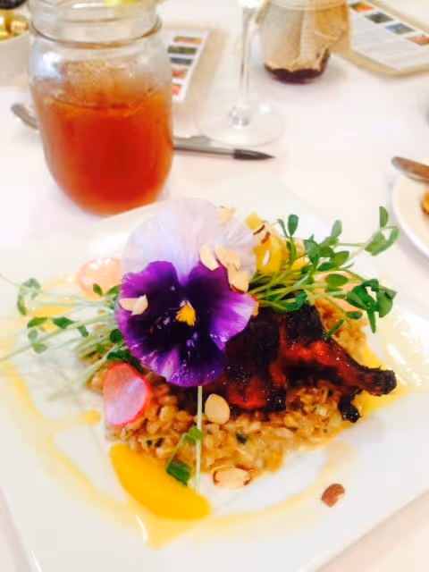 A gourmet plated dish featuring a piece of grilled meat on a bed of risotto or grain, garnished with edible flowers, microgreens, sliced almonds, radish slices, and a peach slice. In the background, there is a glass jar of iced tea with a lemon wedge and a wine glass on a white tablecloth.