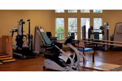 Fitness room with exercise equipment including a recumbent bike, treadmill, and weight machines in front of windows and mirrors.