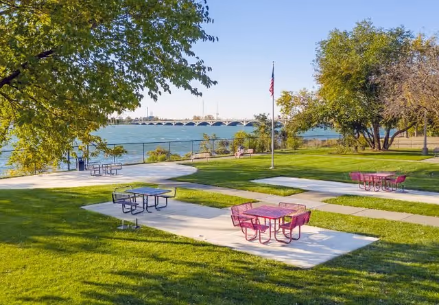 Outdoor park area with green grass, several picnic tables with benches, some painted purple, and a flagpole with an American flag. Trees surround the area, and a body of water with a bridge in the background is visible under a clear blue sky.