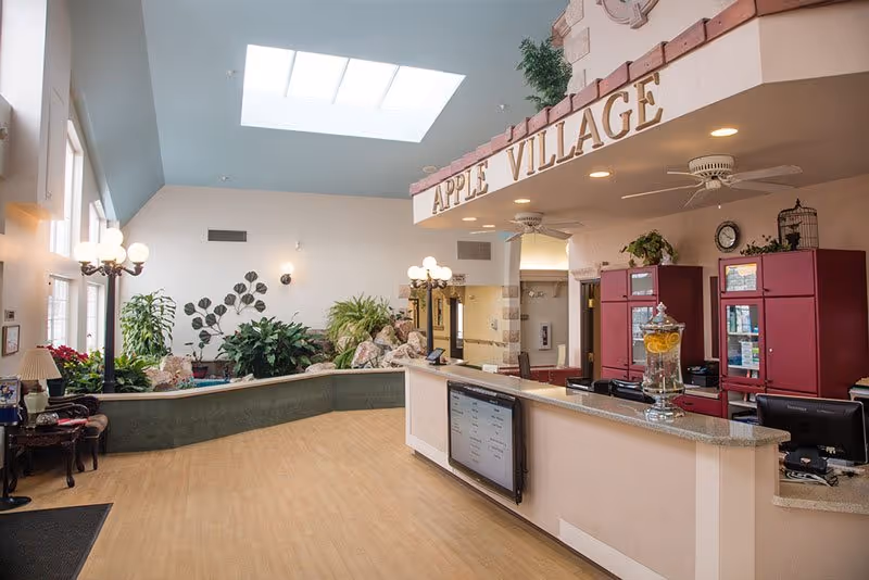 Bright and spacious reception area of Apple Inn At Apple Village with a curved front desk, a large skylight, indoor plants, decorative wall art, and warm lighting fixtures.