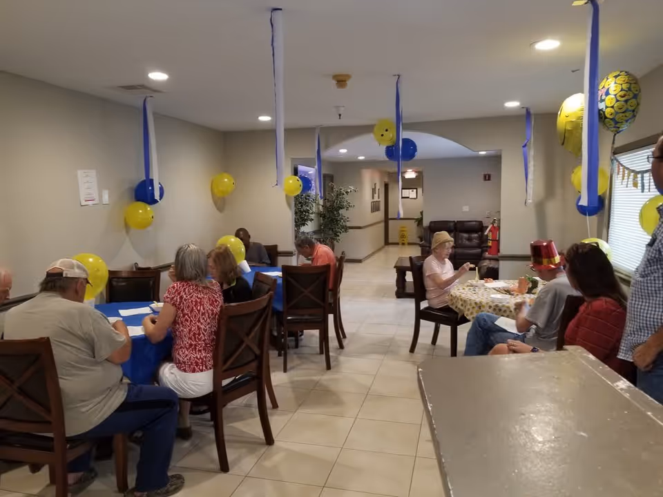 A group of elderly people sitting at tables in a decorated room with blue and yellow balloons and streamers. Some people are engaged in activities or conversation, and the room has tiled floors and neutral-colored walls with overhead lighting.