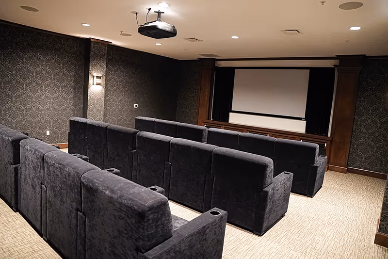 Small screening room with multiple rows of dark upholstered recliner seats, a ceiling projector, and a blank projection screen.