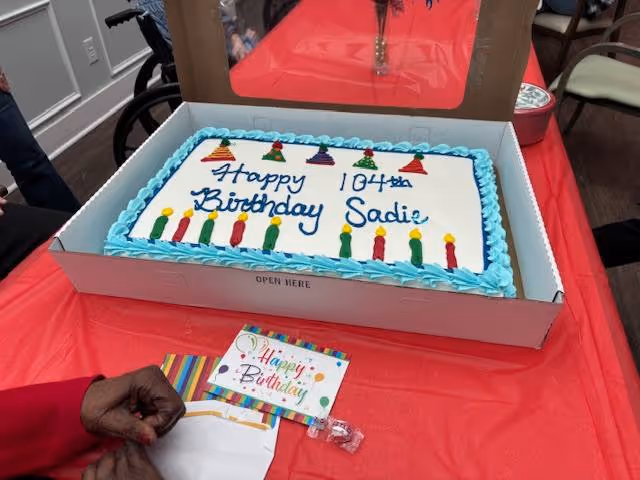 A rectangular birthday sheet cake reading "Happy 104th Birthday Sadie" on a red table with a birthday card and a person's hand nearby.