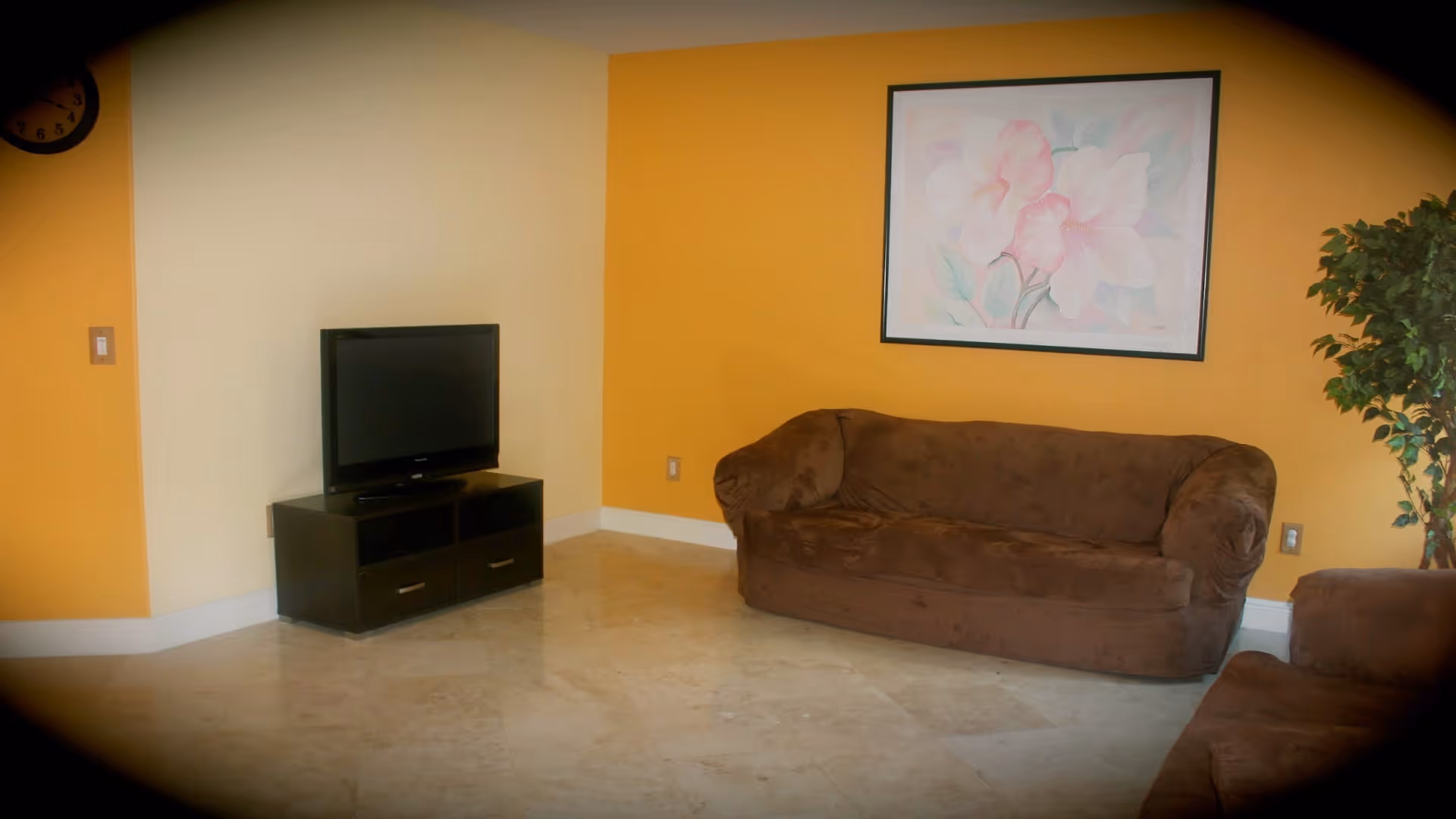 A living room with yellow and beige walls, featuring a brown sofa and loveseat, a flat-screen TV on a black TV stand, a framed floral painting on the wall, a potted plant, and a clock partially visible on the left wall.