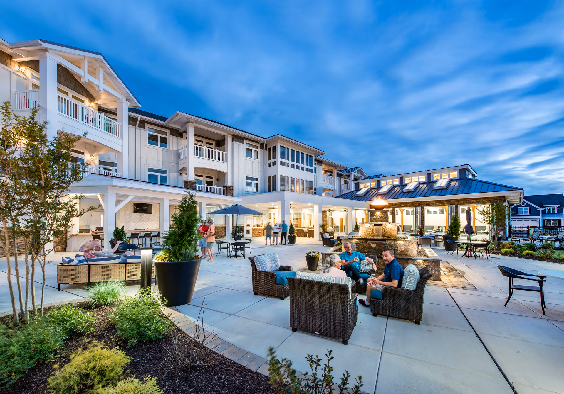 Outdoor patio area of The Lodge at Truitt Homestead during dusk with people sitting and chatting on wicker furniture around a fire fountain. The multi-story building with balconies and large windows is visible in the background under a partly cloudy blue sky.