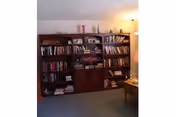 A cozy interior room featuring a large wooden bookshelf filled with books and decorative items. The room has carpeted flooring and a small wooden table with a lamp on the right side.