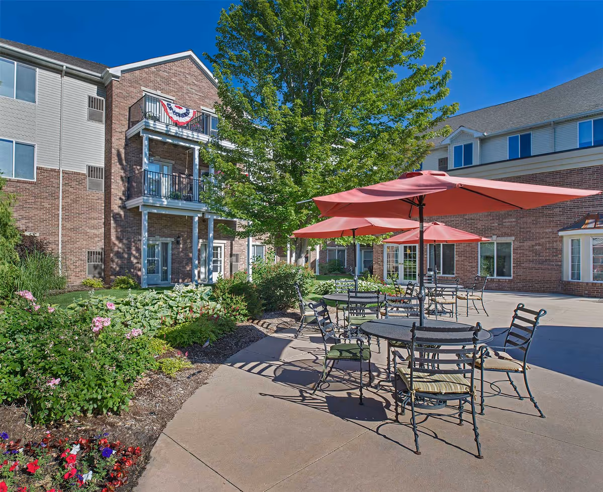 Outdoor patio area at American House Sterling Woods with several round metal tables and chairs under red umbrellas, surrounded by landscaped garden beds and a large tree, with a multi-story brick and siding building in the background under a clear blue sky.
