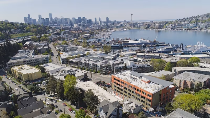 Aerial view of a waterfront urban area with multiple buildings, roads, trees, and a large body of water with boats docked. The city skyline with tall buildings and the Space Needle is visible in the background under a clear sky.
